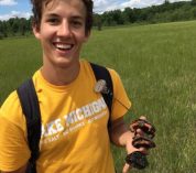 Caleb Krueger stands in a field with trees in the background, holding three small spotted turtles in his hand.