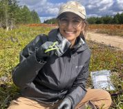 Hannah Rothkopf sits on the ground in a remote area near a dirt road, holding a small green snake.