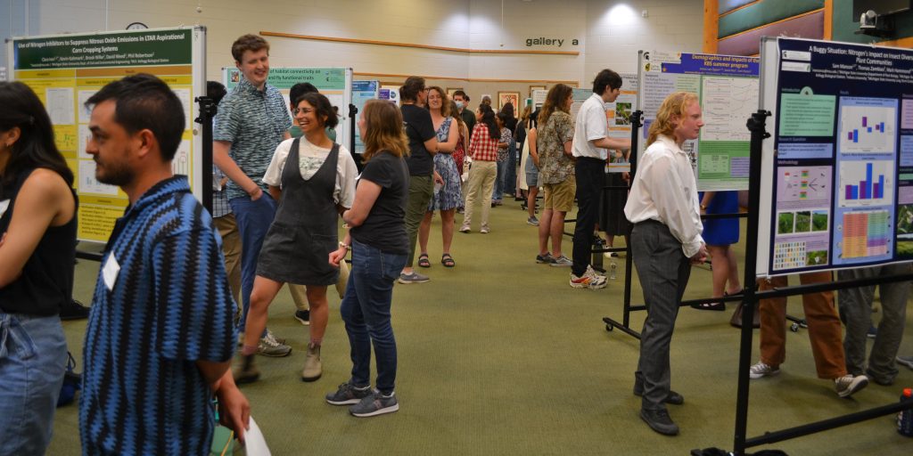 Students and guests gather around large posters mounted on stands at the 2025 W.K. Kellogg Biological Station Undergraduate Research Symposium.