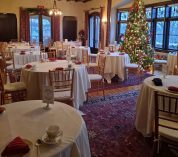 The living room of the W.K. Kellogg Manor House is decorated with a Christmas tree and set for a high tea, with several round tables covered with white tablecloths and place settings.