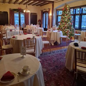 The living room of the W.K. Kellogg Manor House is decorated with a Christmas tree and set for a high tea, with several round tables covered with white tablecloths and place settings.