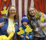 Three "Ducky Day" attendees pose for a photo holding rubber duck props in front of a red-and-white backdrop.