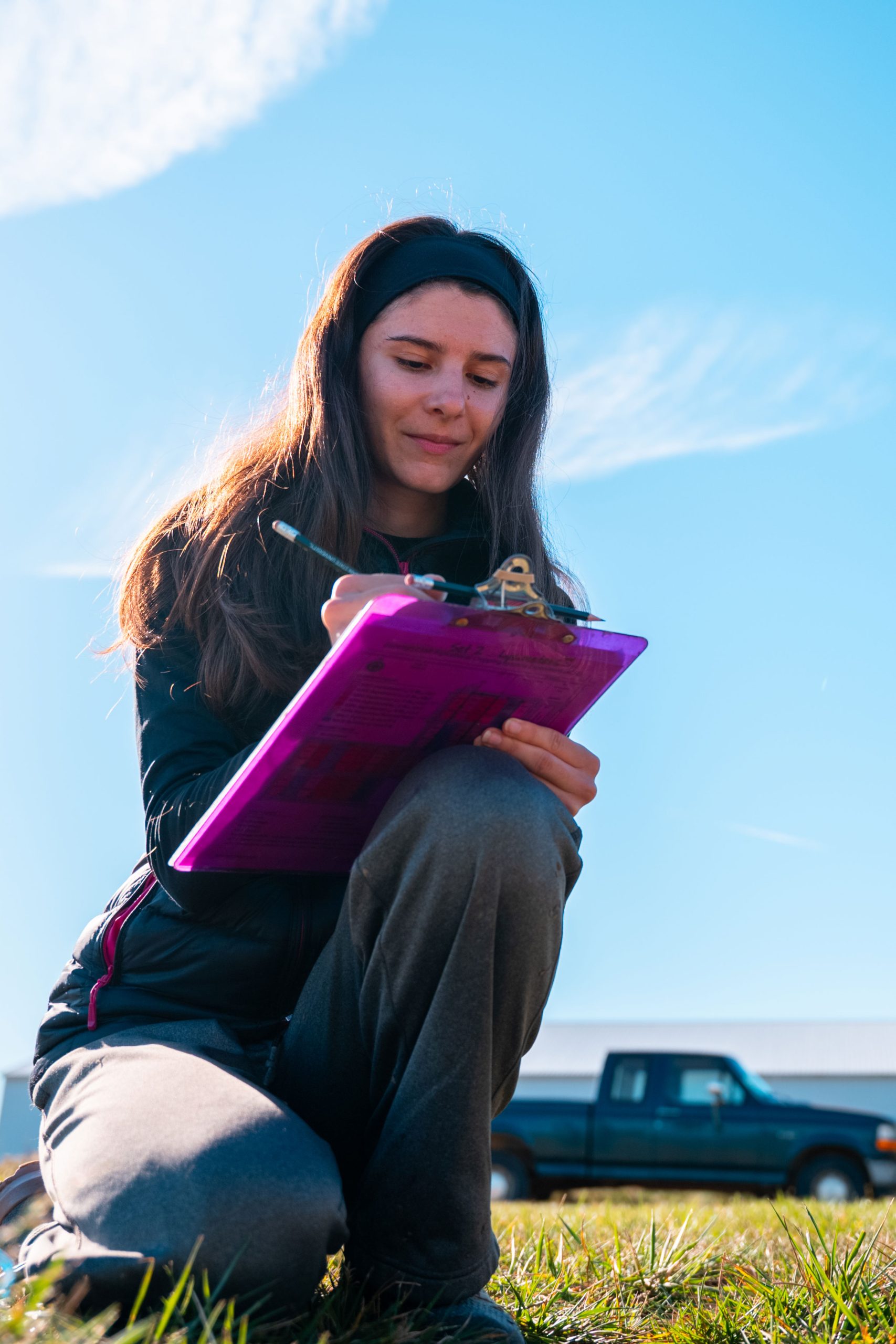 A researcher crouches in a grassy area while taking notes on a clipboard.