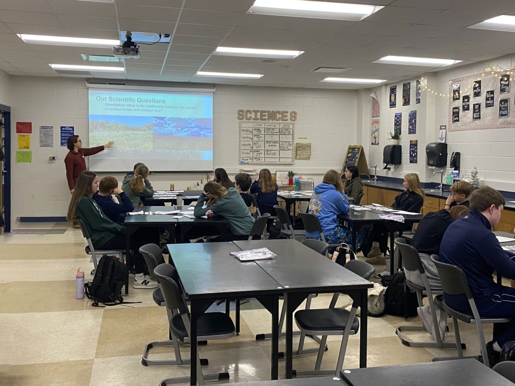 KBS science education and outreach fellow Caleb Krueger gestures to information on a projector screen, with a group of middle school students seated in the foreground.