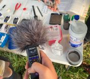 A person sits at a table containing supplies for gathering data, while another holds a sound recording device.